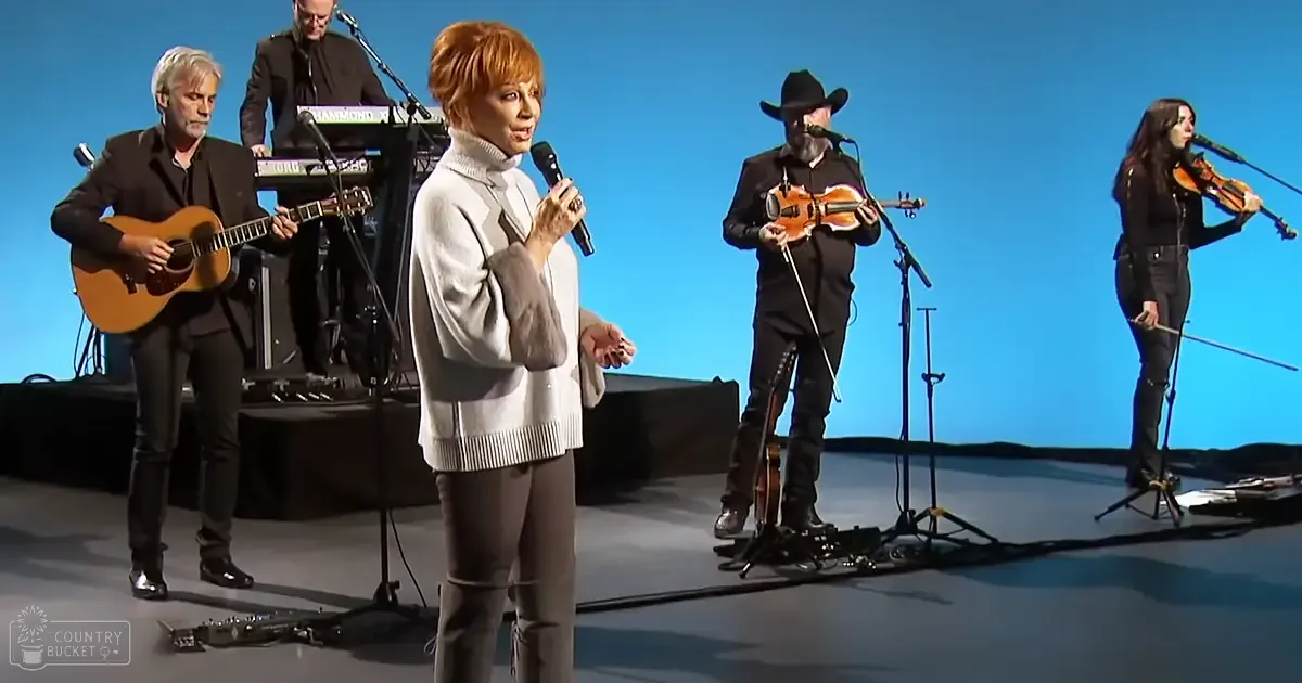 A woman singing on stage with a group of musicians, dressed in casual attire, under controlled lighting.