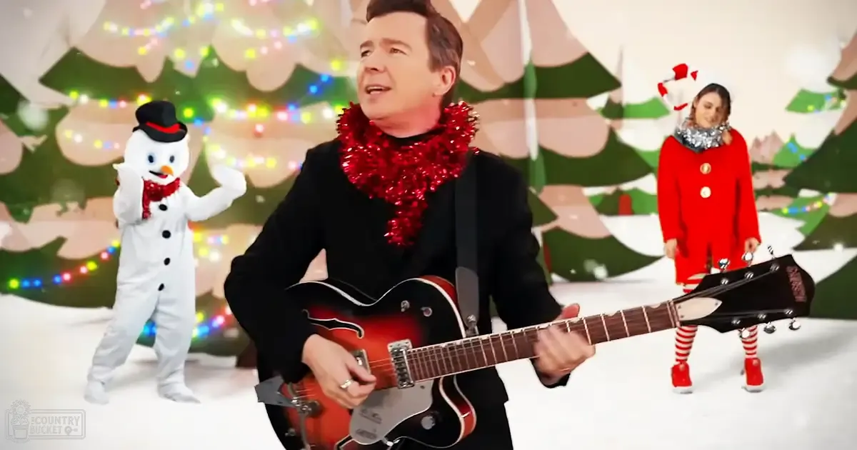 A middle-aged man plays guitar amidst a snowy holiday-themed setting with a child dressed as a snowman, and another woman in a festive red coat nearby.