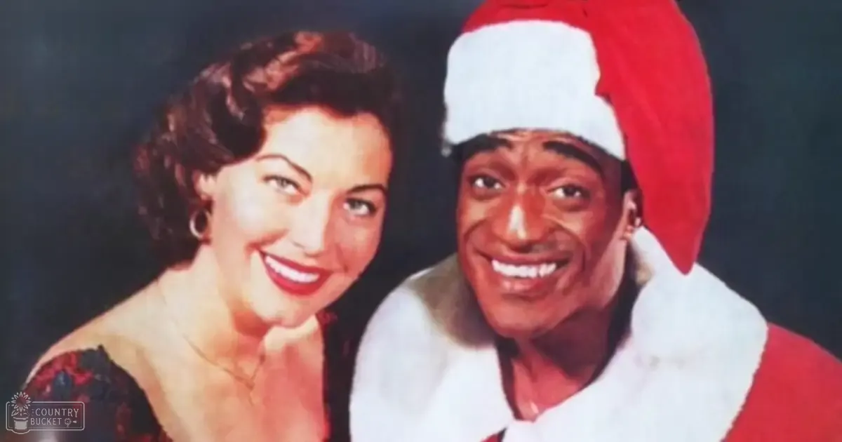 A couple, a female wearing a dark floral dress and an adult male in a Santa Claus costume, posing against a plain studio backdrop during what appears to be a holiday-themed photo shoot.