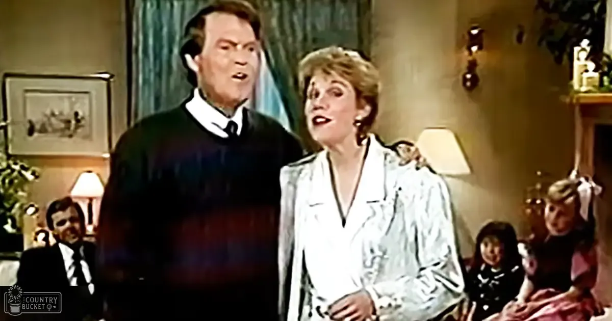 Middle-aged man and woman indoors, man wearing dark striped sweater over white shirt with tie; woman in light V-neck blouse or dress. Framed picture on wall, lamp behind woman, small decorative item on table.
