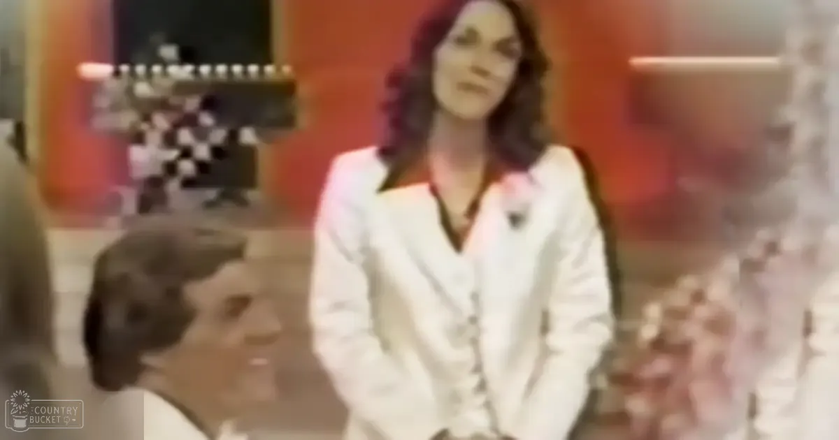 An adult female wearing a white blazer over a collared shirt in an indoor studio or office setting with professional-grade camera equipment visible behind her.