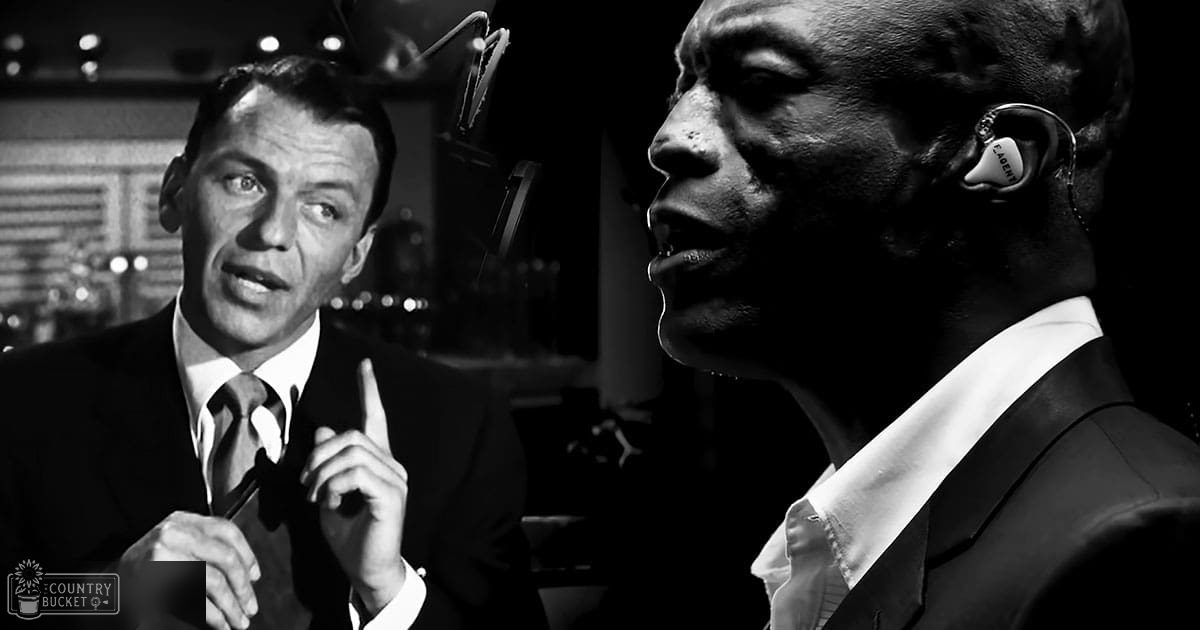 An adult male singer wearing a dark suit jacket with a patterned silk tie in an early to mid-20th century style, seated at a bar or similar venue; another adult male wearing a modern dark suit and earpiece labeled 'EAGEN', indoors with artificial lighting.