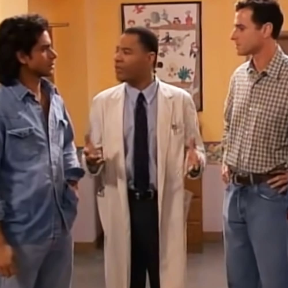 A middle-aged man in a white lab coat stands indoors, possibly in a medical office; next to him is another person wearing light blue coveralls with pockets, buttoned up to the neck. A third individual wears a checkered shirt with light brown and white squares.