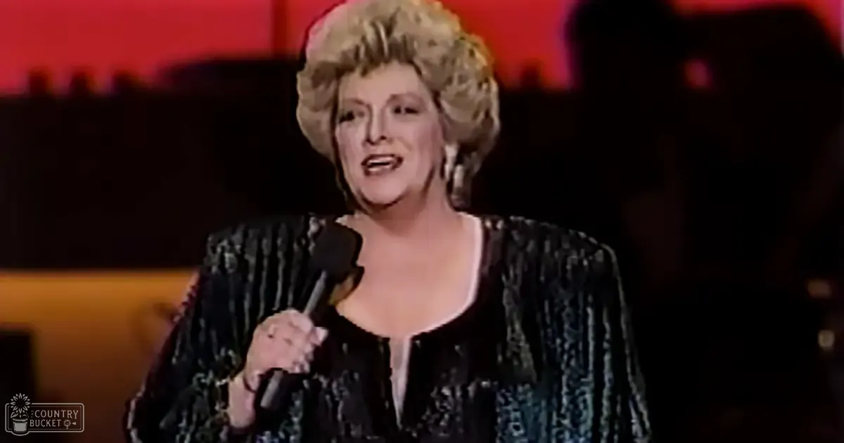 A middle-aged woman wearing a black sequined or glittery jacket with wide sleeves, singing on stage under dim red lighting.