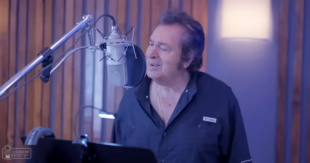 A middle-aged adult male in a recording studio, wearing a dark blue button-up shirt with relaxed fit and collar, surrounded by professional microphone equipment and sheet music.