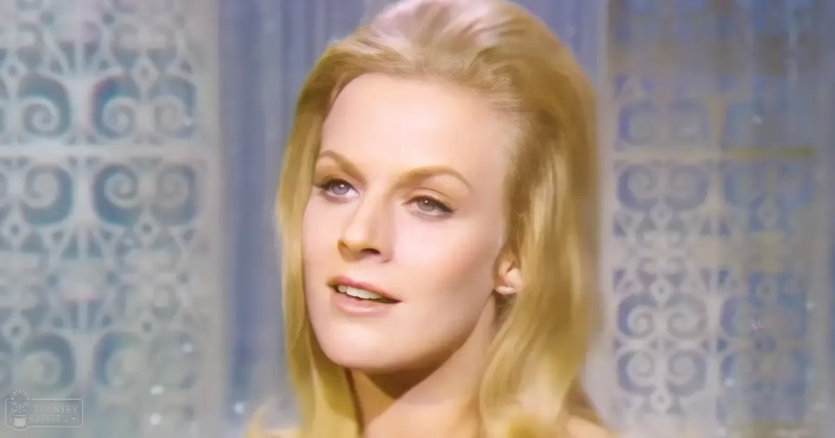 An adult female wearing a fitted, light yellow or beige blouse with a low neckline and smooth fabric indoors, ornate patterned wall in the background.