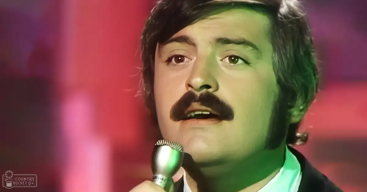 Adult male performer in a formal suit, set against a stage with red lighting and microphone, suggesting an indoor performance or event.