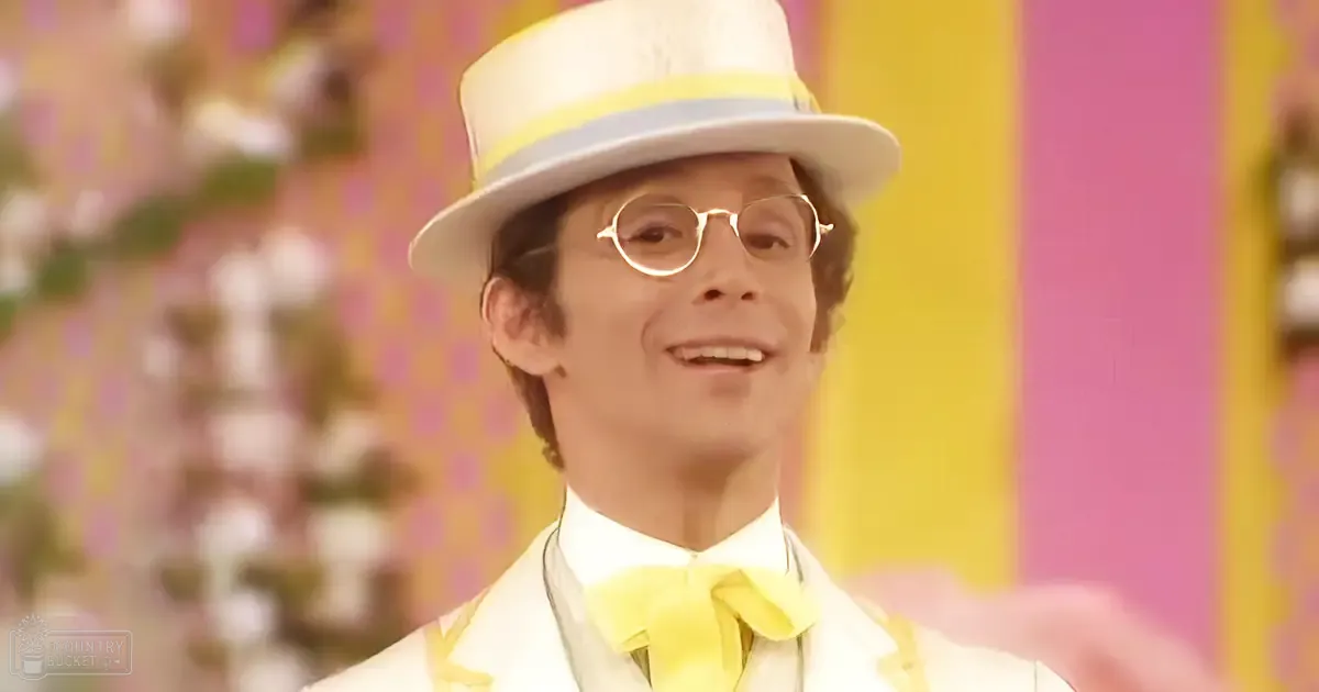 A young adult male in a formal white suit with yellow bow tie and top hat stands on an indoor stage decorated with blurred floral elements, suggesting a performance or event setting.