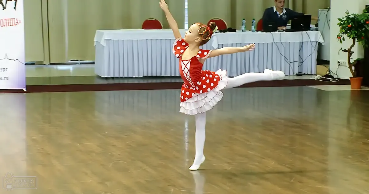 A young female dancer in a red and white polka dot ballet leotard with ruffled skirt, performing in an indoor dance studio setting with bright lighting.