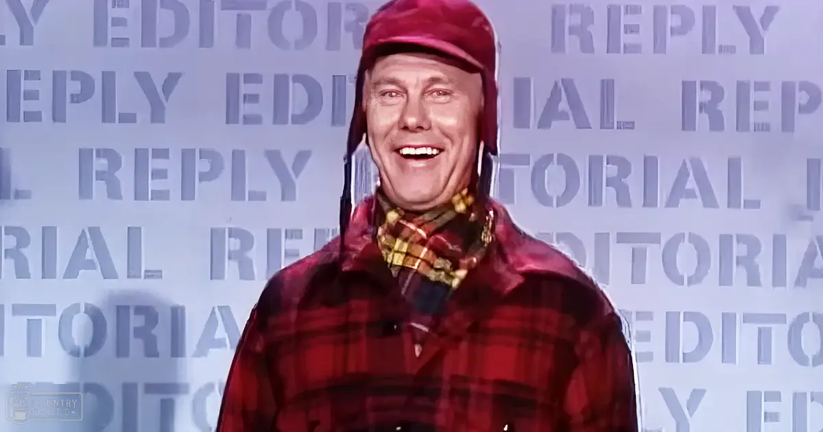 A middle-aged man wearing a red plaid jacket, maroon cap, and yellow checkered scarf stands against an 'EDITORIAL REPLY' backdrop in an indoor studio setting.