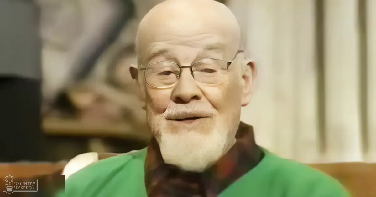 An older adult male wearing a green sweater with a patterned scarf in a softly lit residential setting.