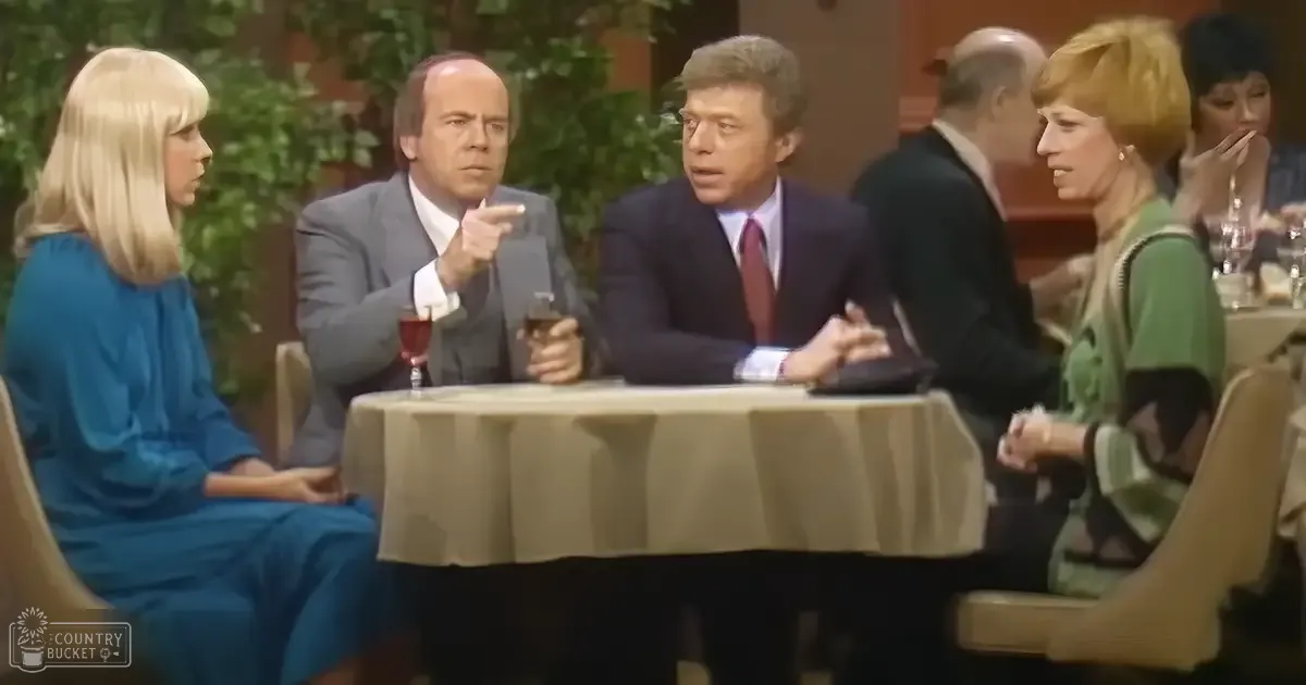A group of four adults engaged in a discussion around a round table covered with a beige tablecloth, set against an indoor backdrop with greenery and bright lighting.