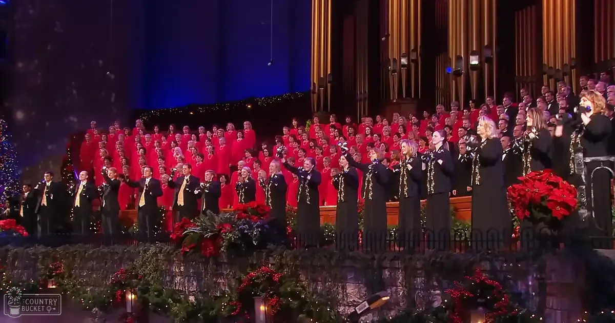 A choir performing in a church or concert hall decorated for Christmas, with adult males wearing dark suits and adult females in black dresses adorned with gold embellishments. The setting is festive with poinsettias and garlands visible.