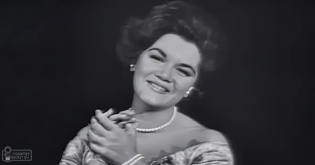 An adult female wearing pearl-like earrings and a long necklace in an indoor setting with dark lighting.