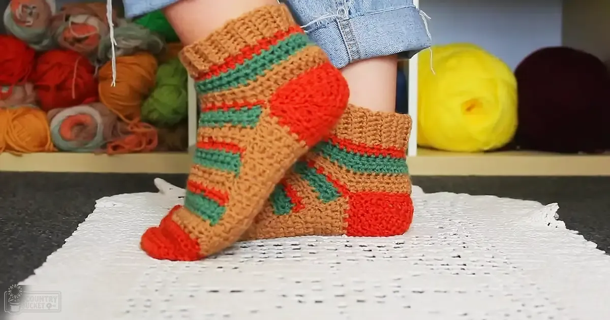 Crocheted socks with orange, green, and brown stripes on a white crocheted doily in an indoor crafting or home setting.