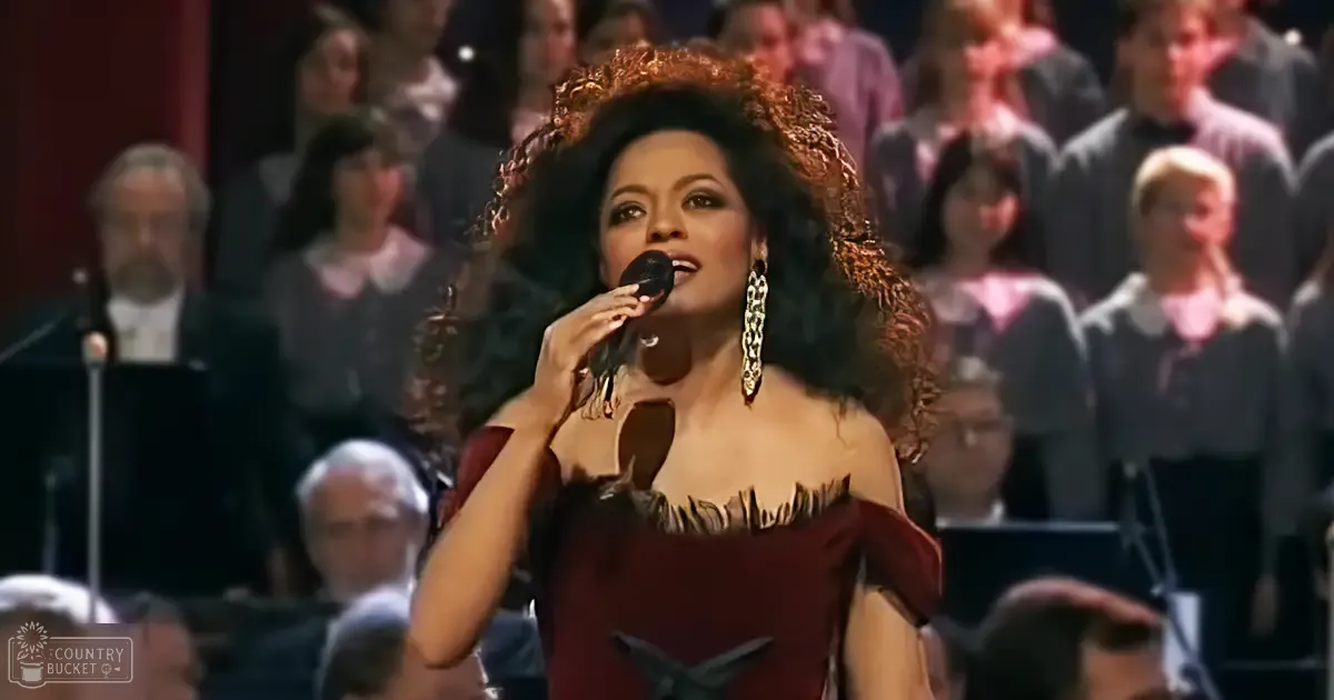 A woman singing in front of an orchestra, wearing a dark red dress with a sweetheart neckline at a concert hall.