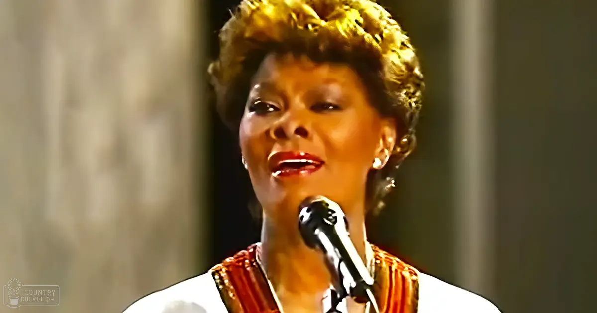 Middle-aged female singer wearing a white garment with red and gold patterned scarf, singing into a black microphone on stage. 'Madly Odd' watermark visible.