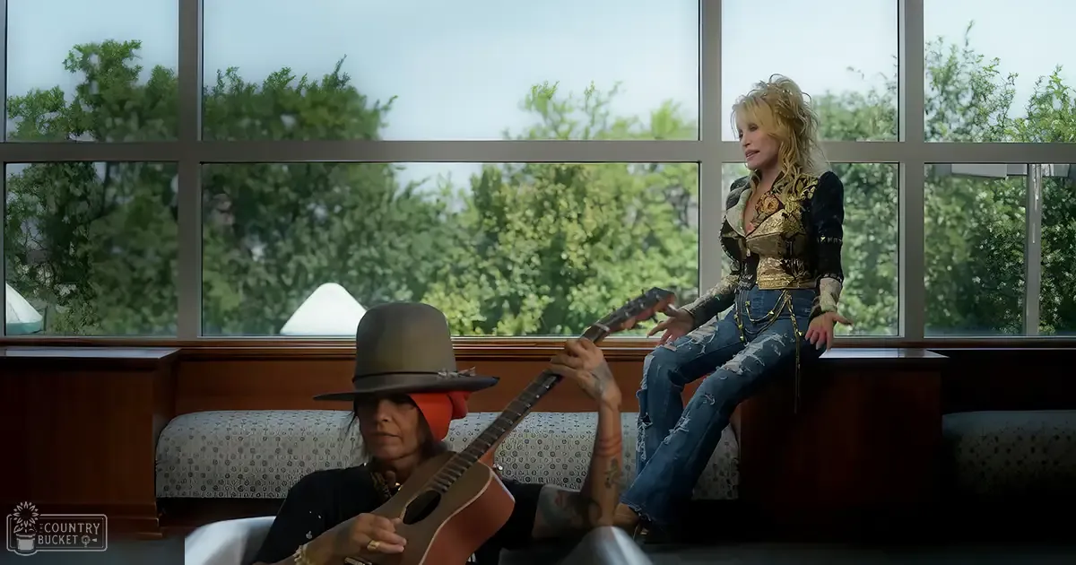 An indoor musical performance with two adults, one wearing a wide-brimmed hat and dark patterned blouse, the other in an intricately designed gold and black top and ripped jeans, surrounded by large windows showing greenery outside.