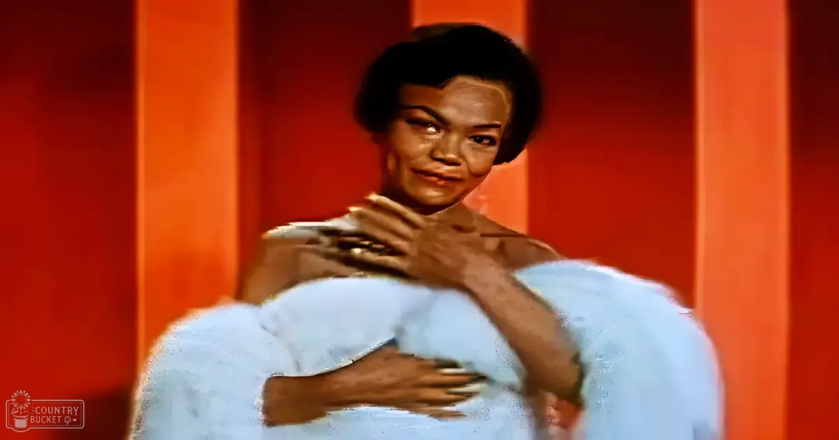 A young adult female in a form-fitting light blue dress with ruffled details, standing indoors near orange walls and vertical panels. A microphone is visible near her mouth.