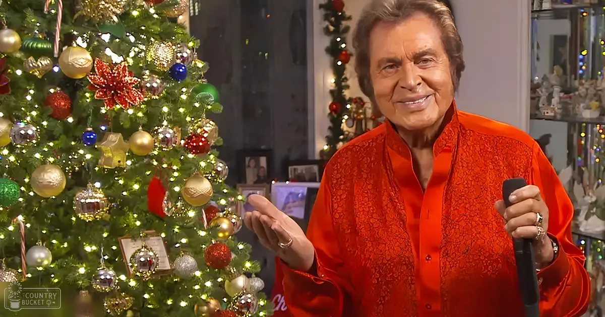 An older adult male in a festive setting, wearing a red lace-patterned shirt and holding a microphone with Christmas decorations around.