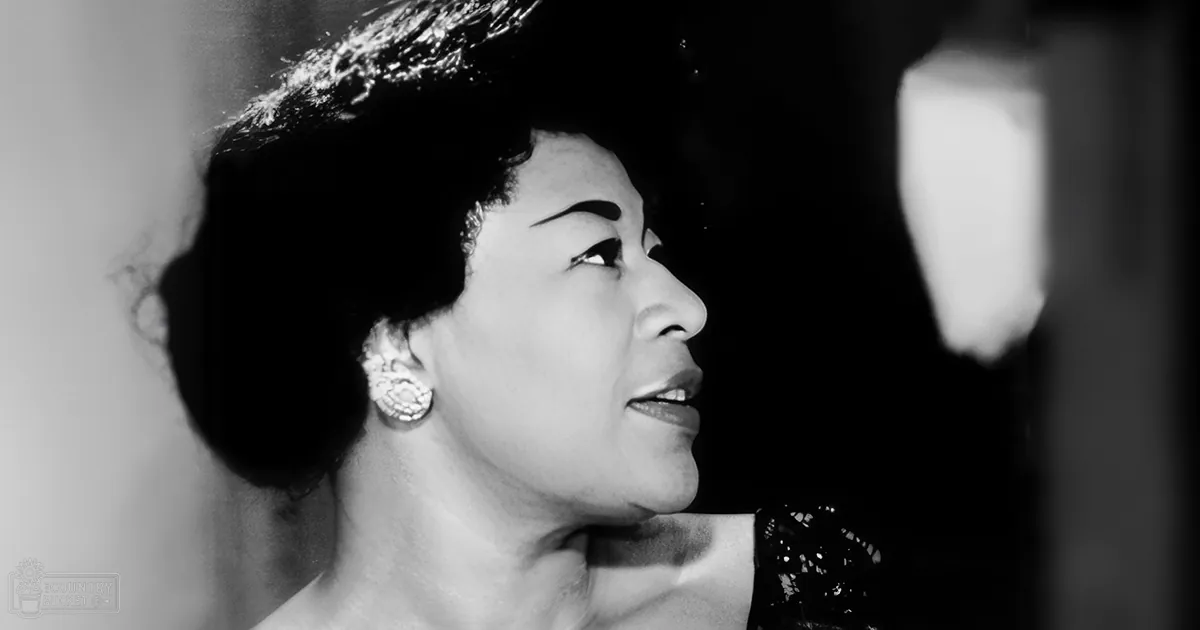 An older adult female wearing a patterned strapless garment indoors, adorned with large earrings, and the background includes part of a curtain or drapery with light reflections.