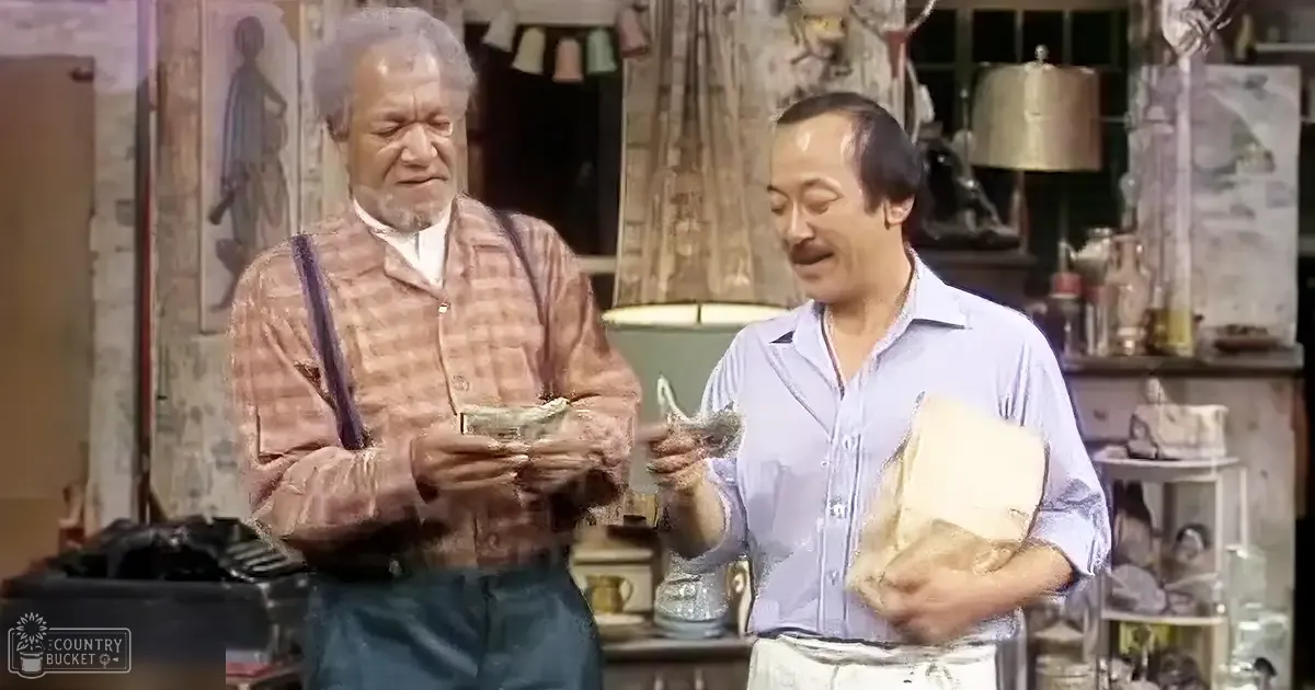Two middle-aged men in an indoor workshop with shelves of sculptures and books, patterned curtain, brown and blue tones.