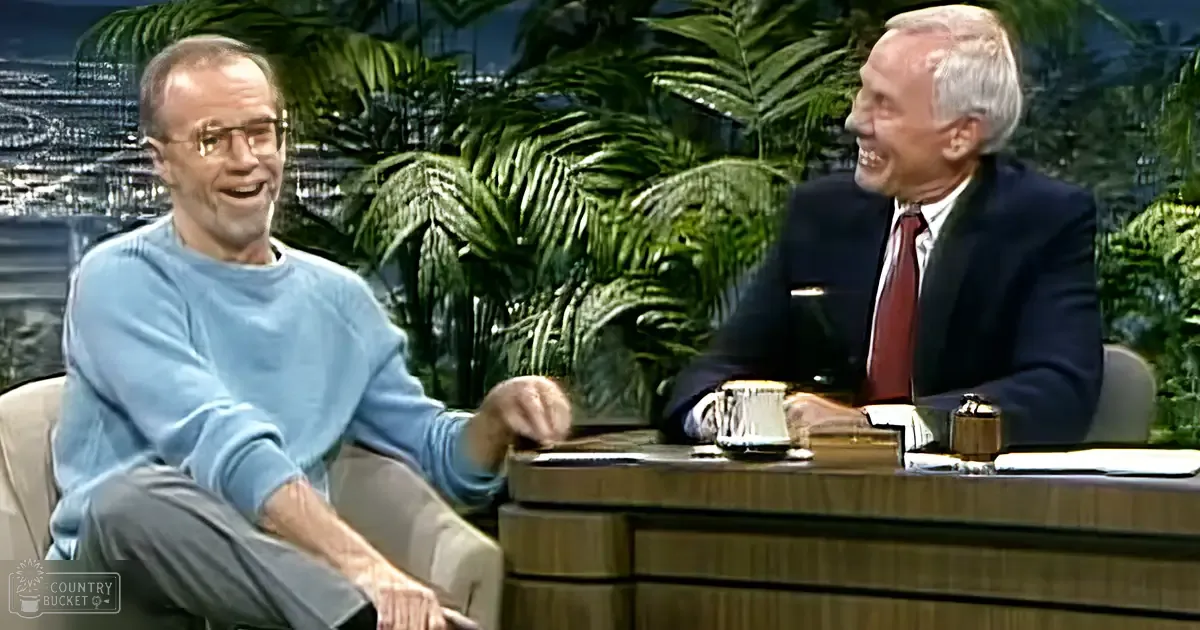 Two older adults conversing in a talk show studio with wooden desk, green plants, and soft-focus cityscape backdrop.