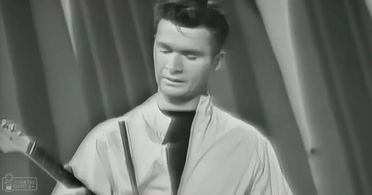A young adult male wearing a white zip-up jacket stands indoors, possibly in a performance venue with vertical curtain folds and evening lighting.