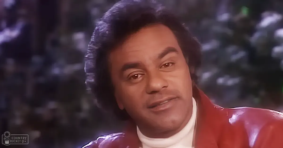 A middle-aged adult male wearing a red leather jacket with a shiny finish, paired with a white turtleneck, stands on what appears to be a stage in an indoor performance venue.