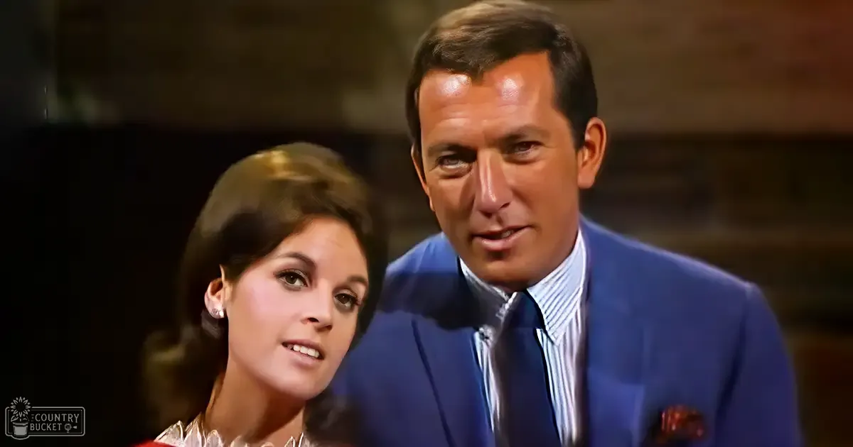 A middle-aged man and a young adult woman in an indoor setting, with the man wearing a blue suit jacket, white shirt, dark tie, and red pocket square; the woman is dressed in a sleeveless floral-patterned top.