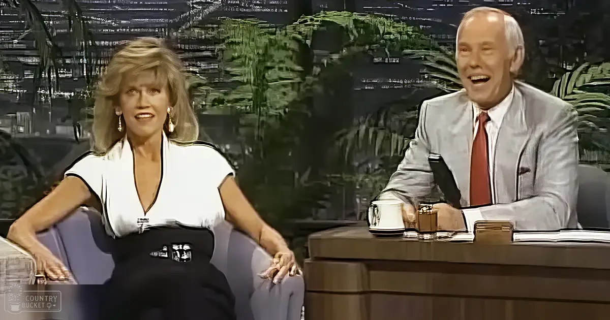 A talk show host and guest in a studio setting with professional lighting and mid-20th-century decor with a desk, mug, abstract foliage background, white blouse with black trim, light gray suit jacket, red tie.