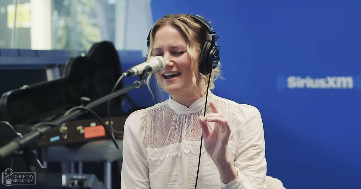 A young adult female wearing a white lace-detailed blouse and black headphones stands at a microphone in a SiriusXM recording studio with 'MADLY ODD' equipment visible behind her.