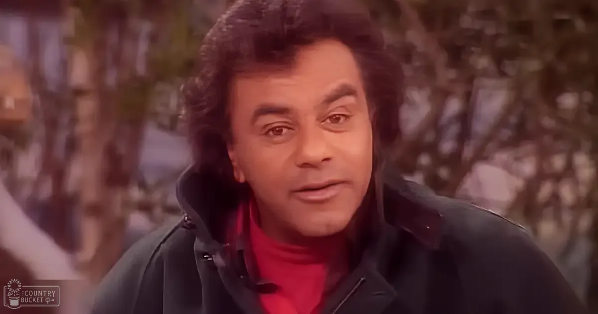 An adult male in a layered outfit, dark jacket over red turtleneck, outdoors with muted natural light and earthy tones.