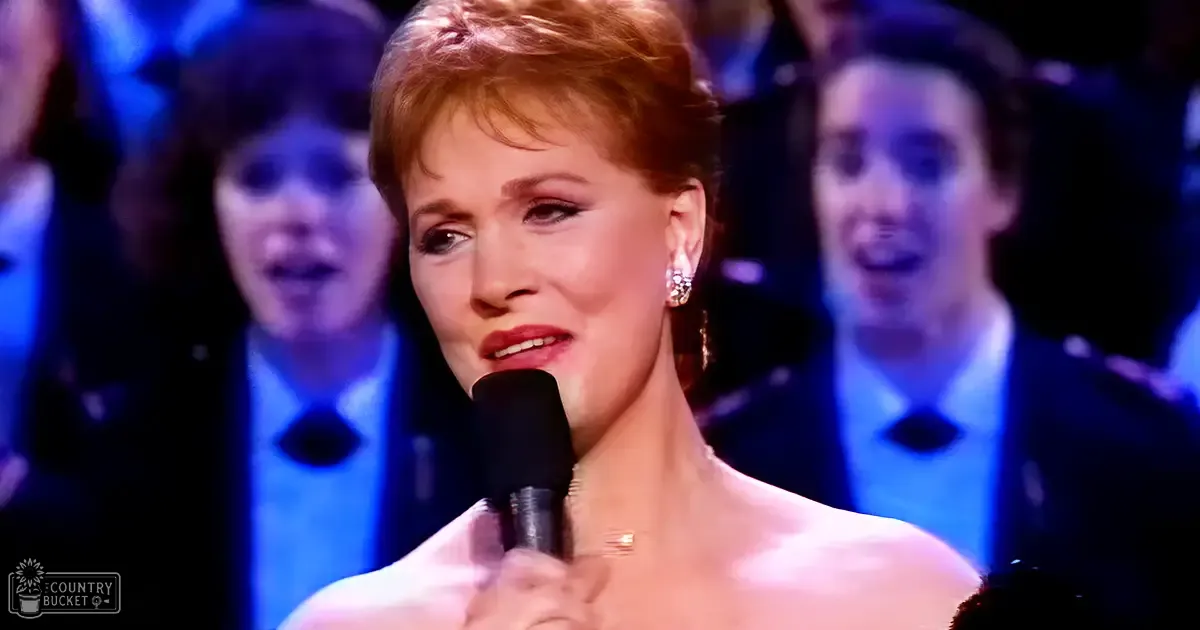 A middle-aged female singer in a strapless dress with a sweetheart neckline and necklace performs on stage at an event, surrounded by formally dressed individuals.