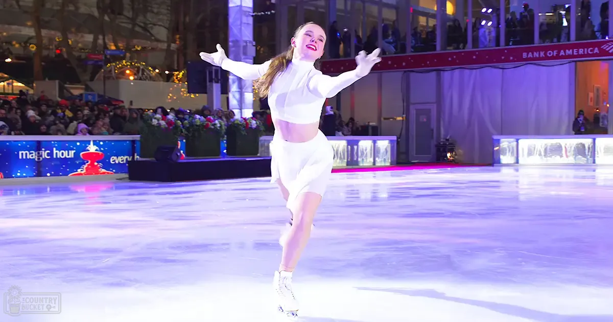 A young adult female figure skating on an outdoor ice rink at Bank of America Winter Village in New York City, surrounded by winter decorations and evening lighting.