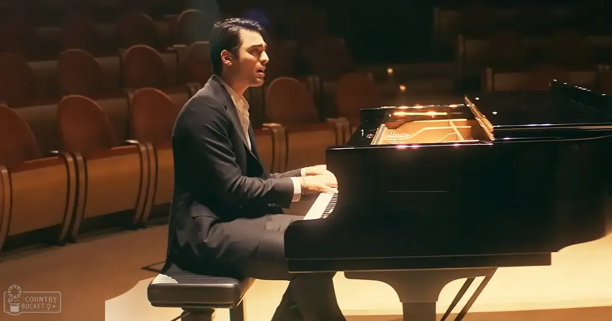Adult male playing piano in an indoor auditorium with warm lighting and rows of empty seats.