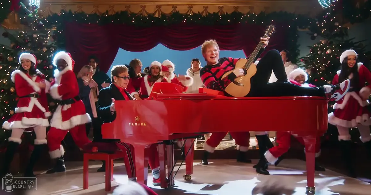 A young adult male plays a guitar in front of a red Yamaha grand piano, surrounded by festive Christmas decorations and other adults dressed for the occasion.