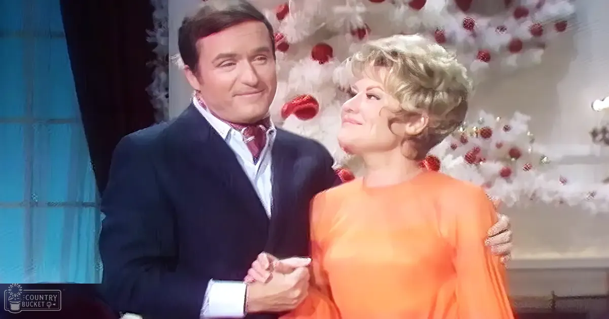 A middle-aged couple dressed festively, with a man wearing a dark suit and white shirt with red patterned tie, and a woman in an elegant orange silk dress, standing indoors against the backdrop of a decorated Christmas tree during daytime winter season.