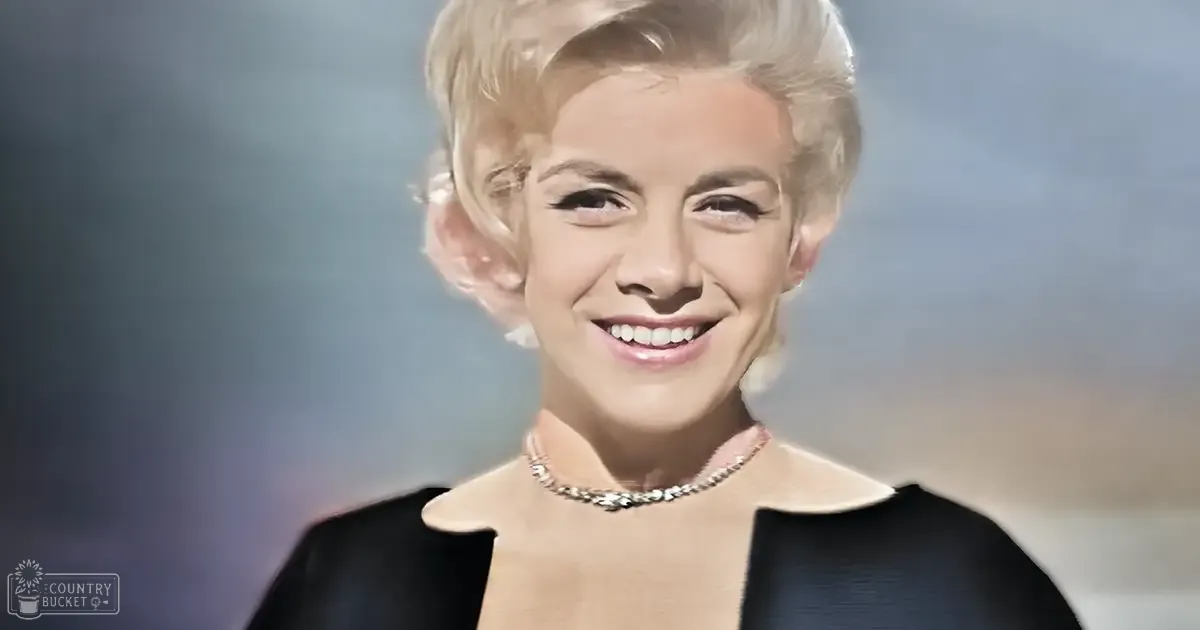 A young adult female wearing a black, form-fitting dress with structured shoulders and a high neckline, accessorized with a detailed necklace, set against a softly lit studio backdrop.