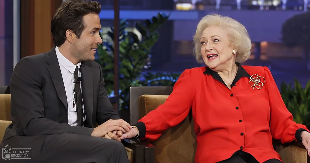 A young adult male guest and an older female talk show host wearing formal attire, seated on a brown couch with plants in the background.