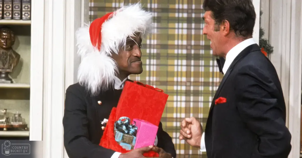 A man wearing a Santa hat with white fur trim holds gifts alongside another middle-aged to older adult male in a black suit and red pocket square, set indoors possibly during Christmas time.
