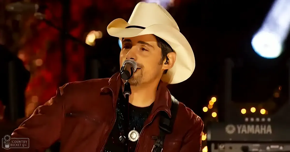 An adult male performer in a dark red leather jacket, black shirt, and white cowboy hat with a black band stands on an indoor concert stage. Blurred lights and stage equipment suggest the setting is during a live music performance.