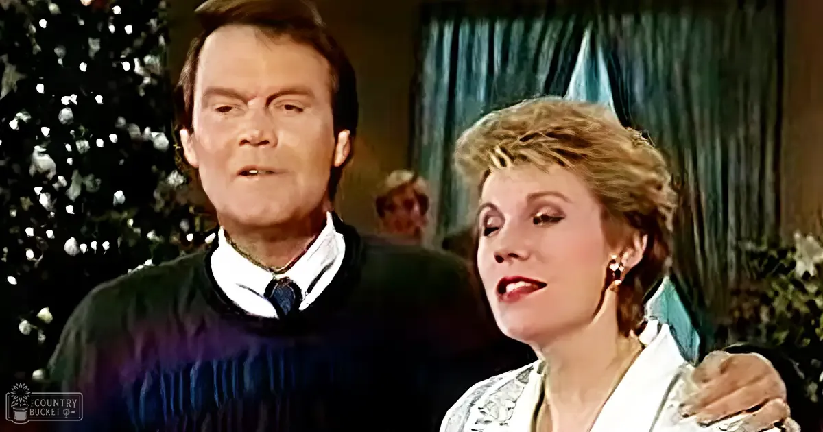 Middle-aged male wearing a dark striped sweater over a white collared shirt with a tie, standing next to a middle-aged female in a white top with intricate detailing and a fur-like accessory draped over her shoulder. A Christmas tree is partially visible on the left.