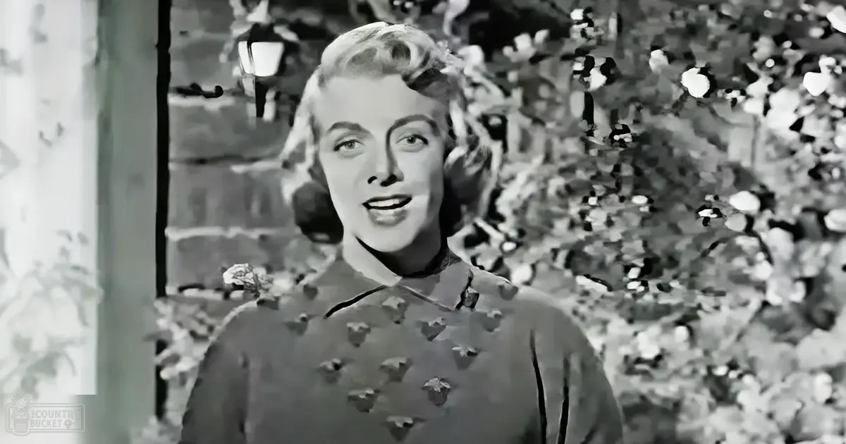 A young adult female in a relaxed, loose-fitting long-sleeved dress with a floral pattern, standing outdoors among lush greenery and flowering bushes under natural daylight.