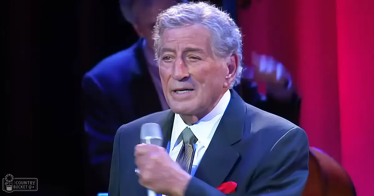 A middle-aged man in a dark suit with a white shirt and striped tie, holding a microphone against a backdrop of red curtains and dim lighting, possibly on stage.