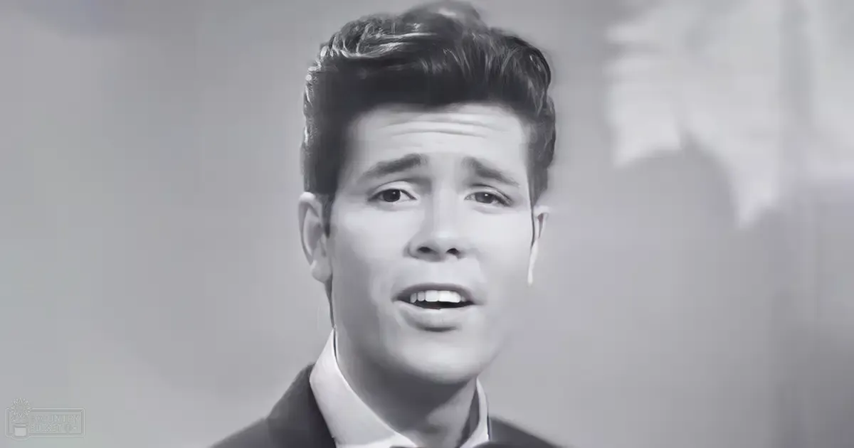 Young adult male in a studio setting, wearing a dark jacket over a crisp white shirt with tailored sleeves, monochromatic photograph.
