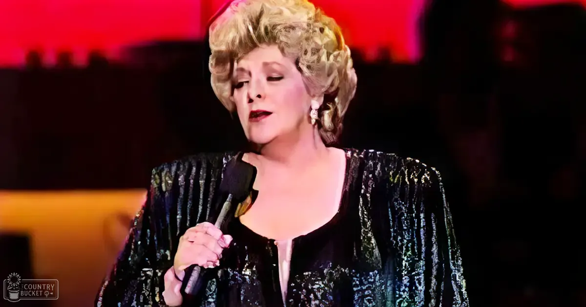 A middle-aged female performer in a black sequined jacket with pearl earrings, holding a microphone on stage under red lighting.