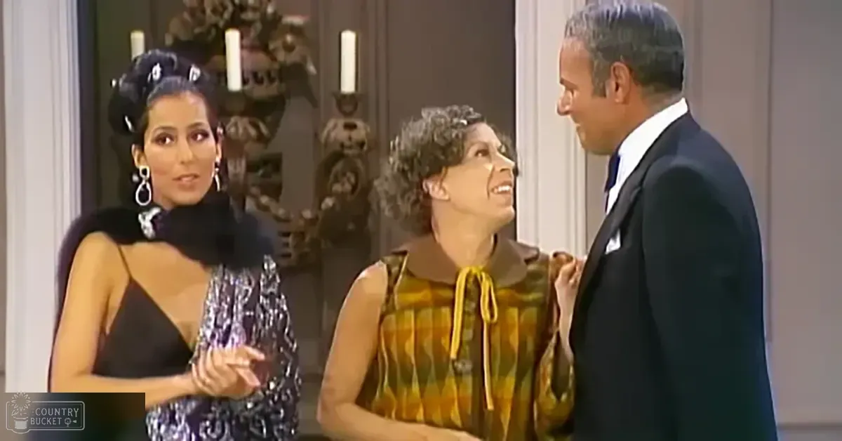 A woman wearing a black dress with sequined bodice and fur collar stands next to another woman in a yellow patterned top, both dressed formally for what appears to be a dinner party. A man in a black suit completes the group.