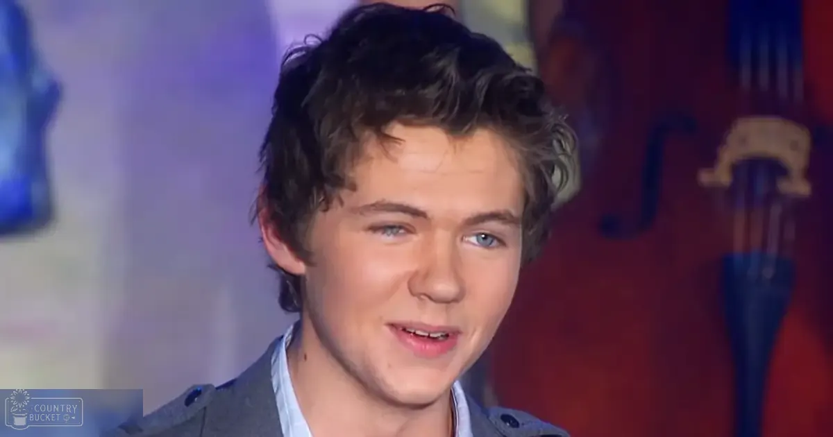 A young adult male wearing a relaxed fit button-up shirt with long sleeves, light blue color, over a dark gray or black jacket, stands indoors near what appears to be part of a musical instrument, possibly a cello or double bass. The setting suggests an indoor stage or performance area.