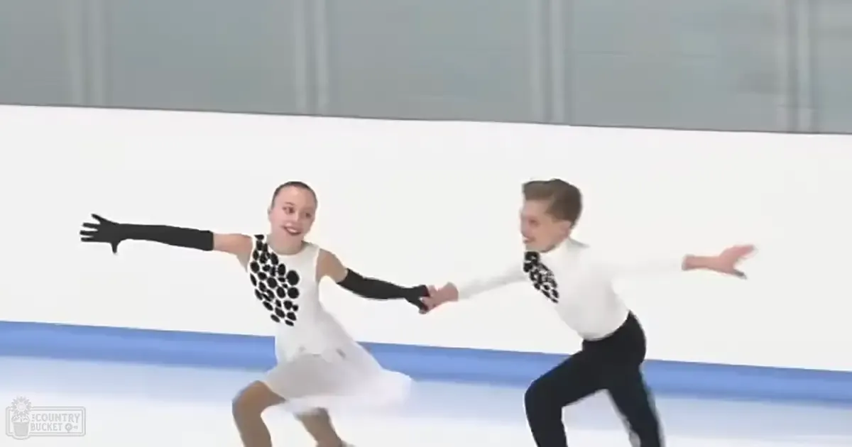 A figure skating pair in white dresses with black polka dots, performing indoors under artificial lighting on an ice rink.